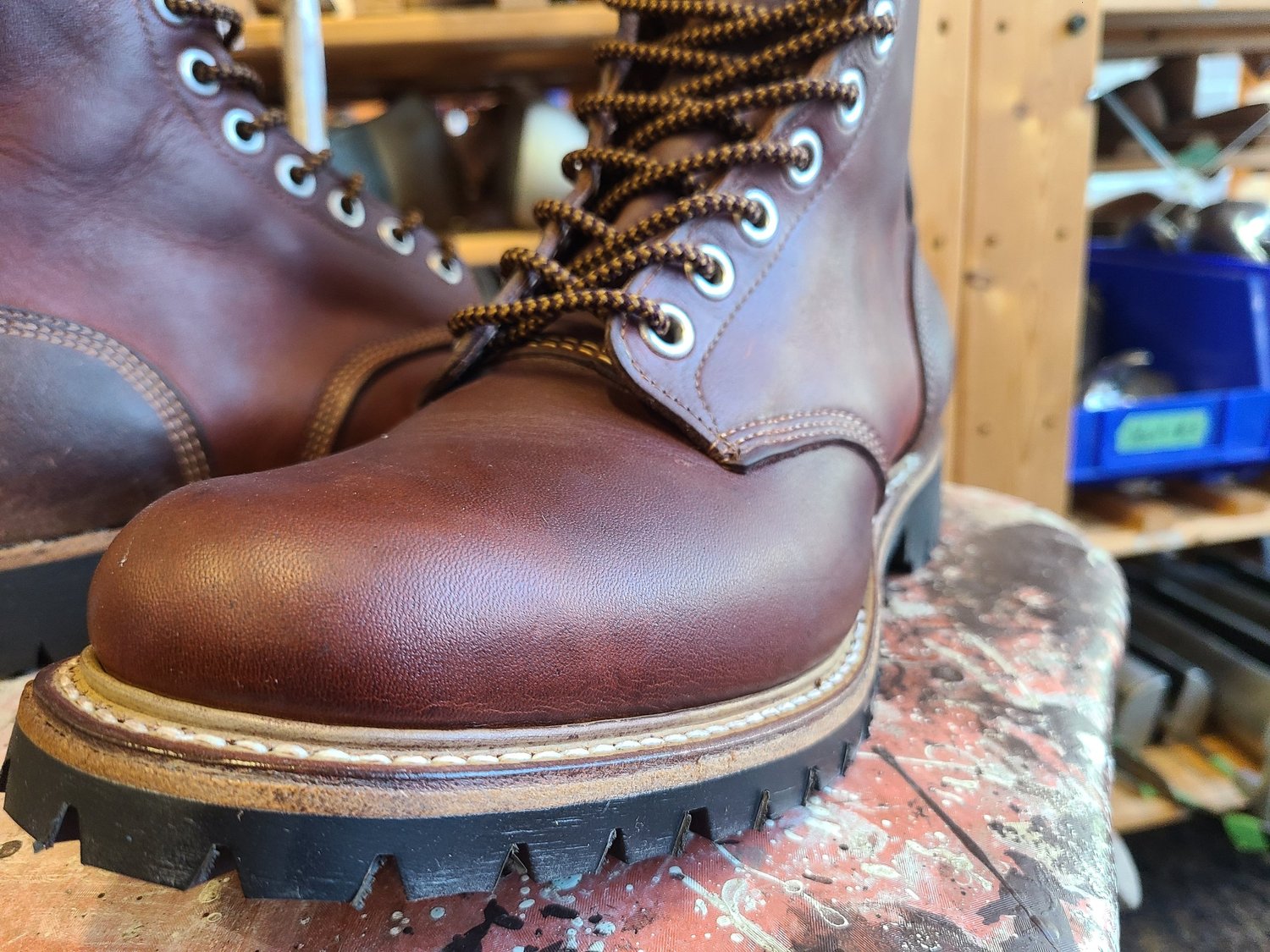 A pair of brown leather work boots with black rubber soles and yellow-brown laces on a wooden surface in a workshop setting.