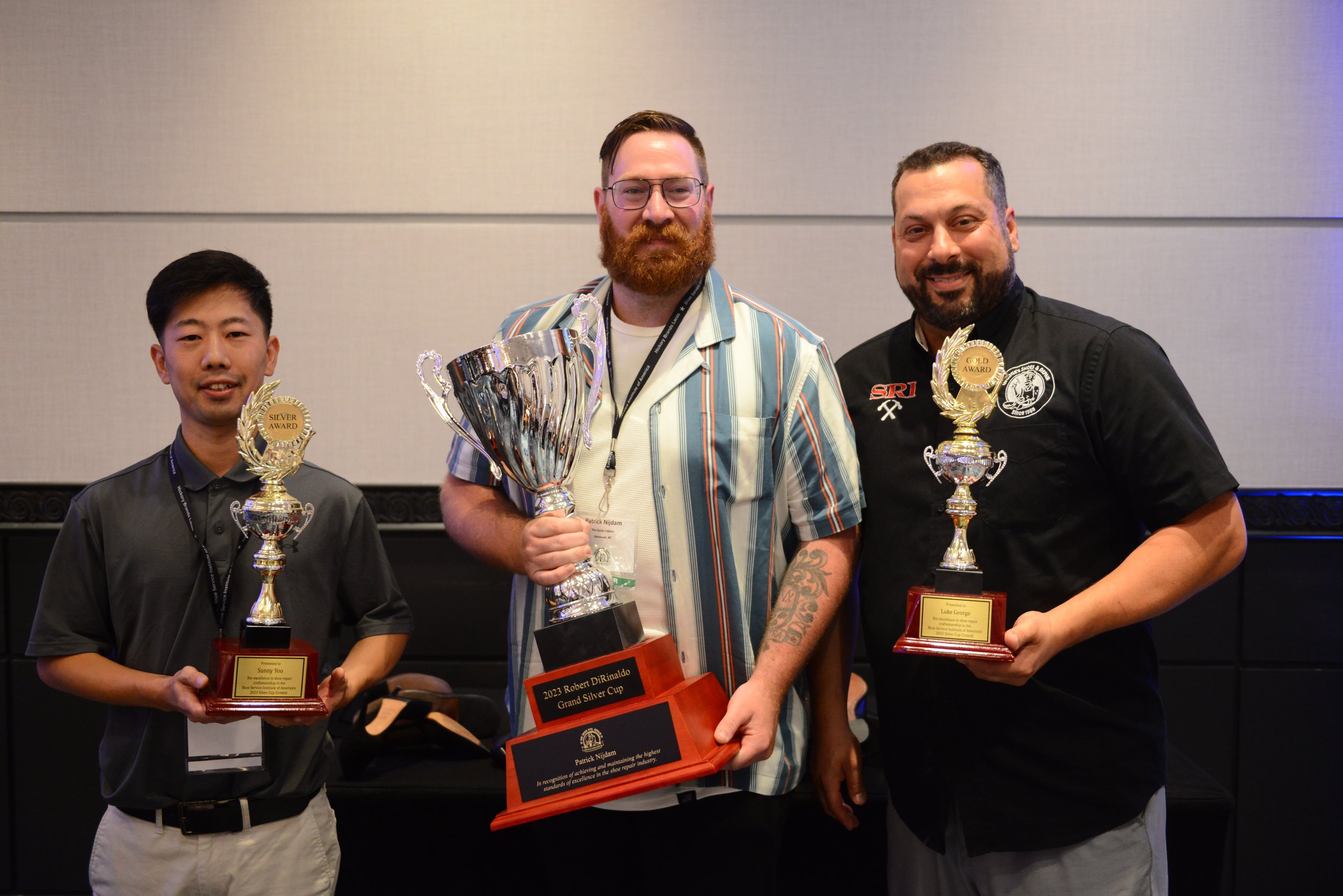 Three men standing together holding trophies, celebrating an achievement at an indoor event. The middle man has a large silver cup and a plaque that reads '2023 Robert DiRinaldo Grand Silver Cup.'