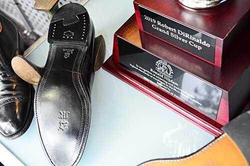 A black dress shoe, a wooden plaque or trophy labeled '2019 Robert DiMald Grand Silver Cup', and part of a brown shoe are shown on a glass surface.