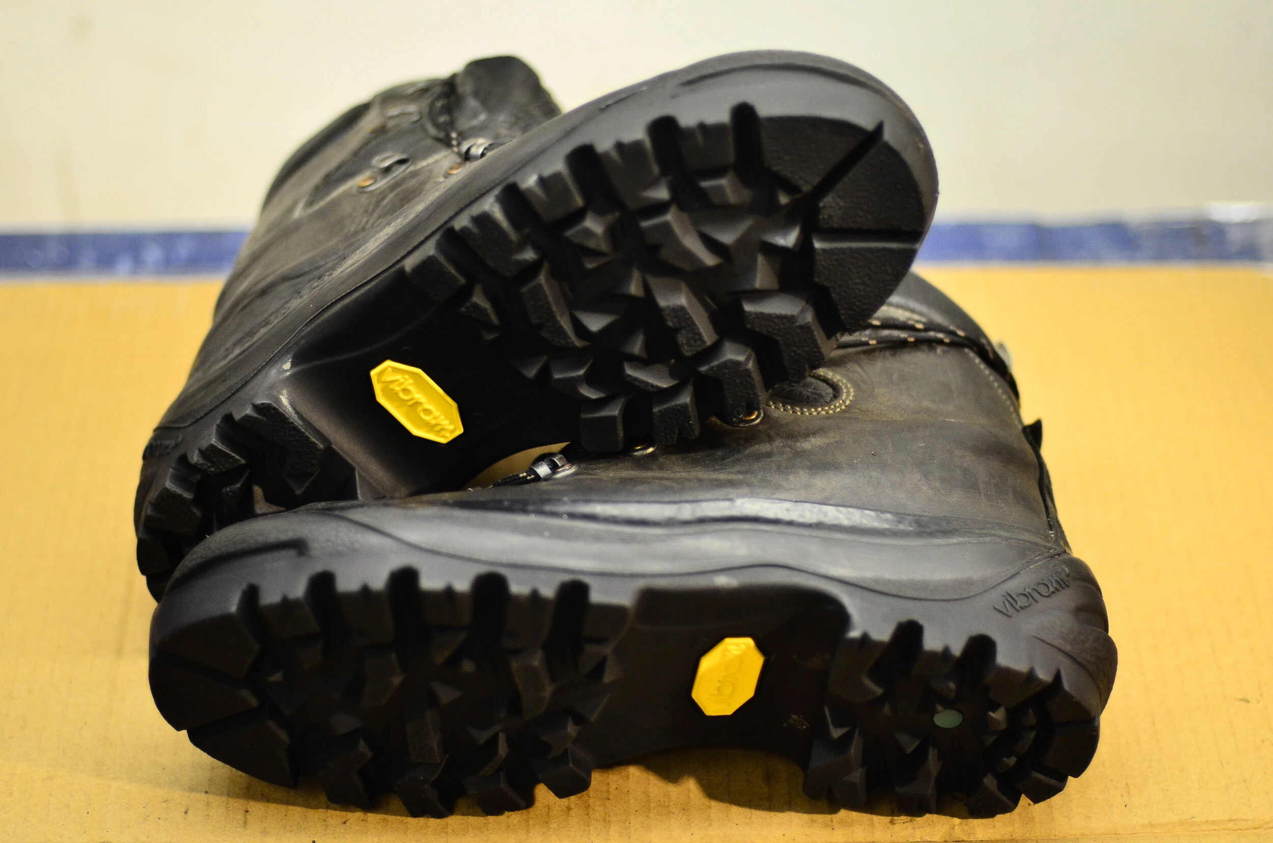 A pair of black hiking boots with Vibram soles and yellow Vibram labels on the bottom, resting on a cardboard surface.