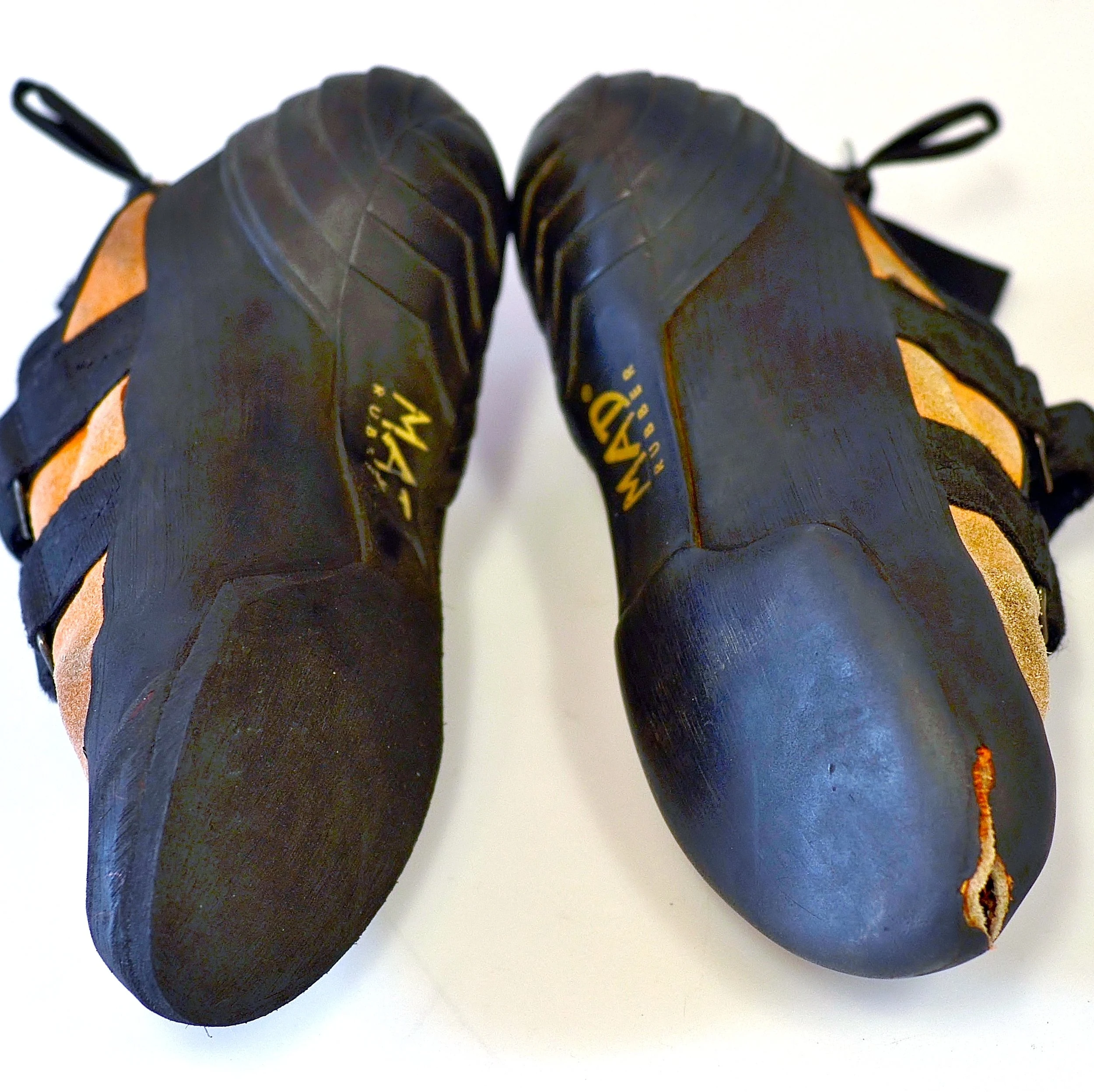 A pair of black and orange climbing shoes with visible wear, labeled 'Mad Rock', placed on a light-colored surface.