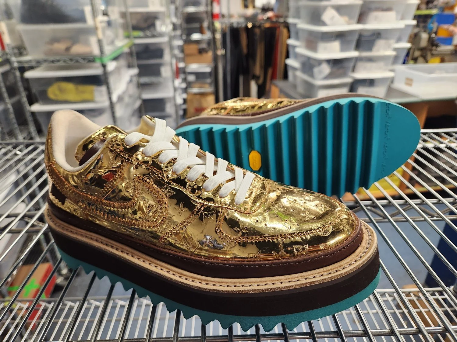 A gold-colored sneaker with decorative chains and detailed embroidery, placed on a metal rack in a storage or warehouse area.