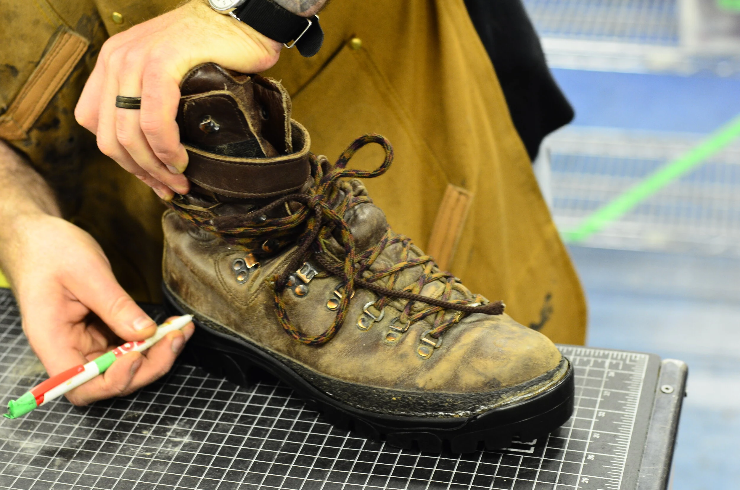 A person working on a worn, brown leather hiking boot that is placed on a cutting mat, with a pen in hand and a bright workshop background.